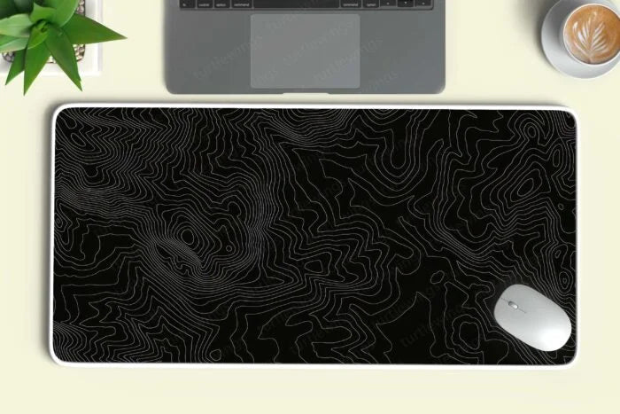 Large black custom desk mat with white topographic contour lines next to a laptop, mouse, plant, and coffee cup